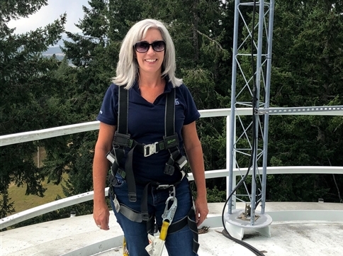 A white woman stands on top of a generation tower strapped into a safety harness overlooking trees