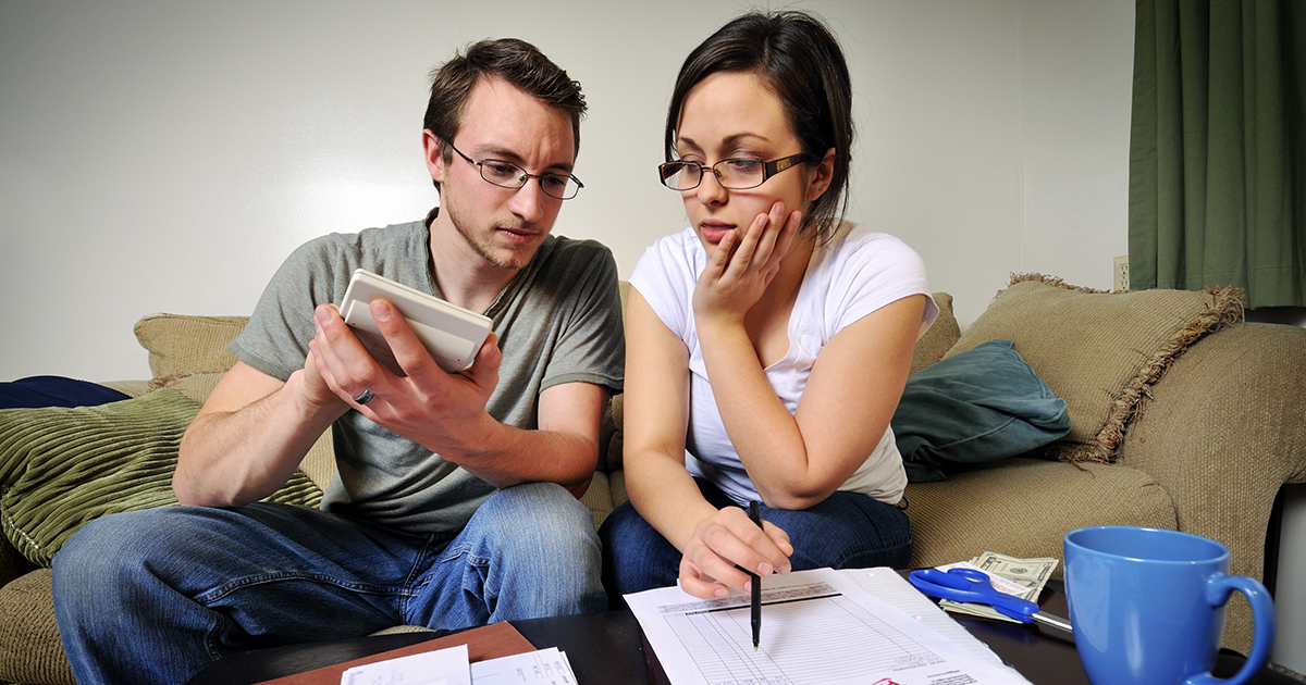 Man and woman sitting on a couch, looking at bills with pens and calculators in hand