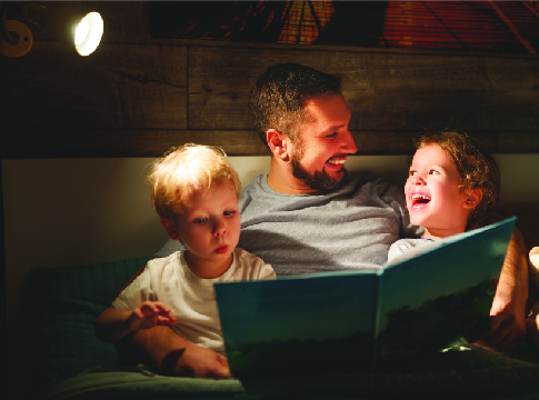 father with 2 young kids, reading a book by lamplight