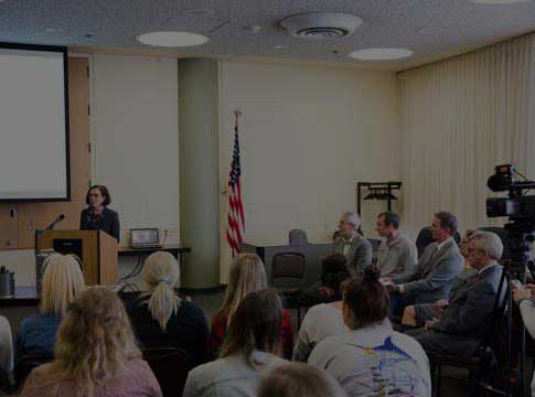 Oregon Governor Kate Brown standing at a podium in room full of seated people.