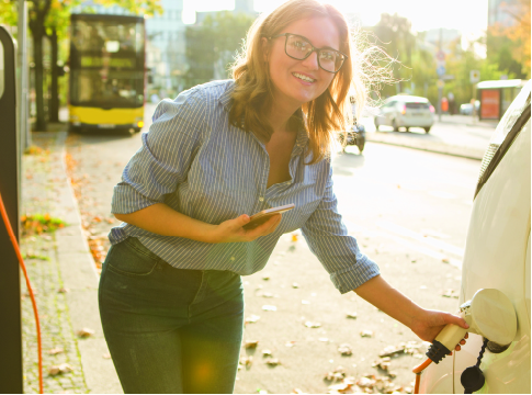 Smiling young woman with glasses holding her cell phone and plugging in her EV