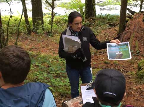 Woman wearing jacket with EWEB logo showing a picture to a group of students with the McKenzie River in the background.
