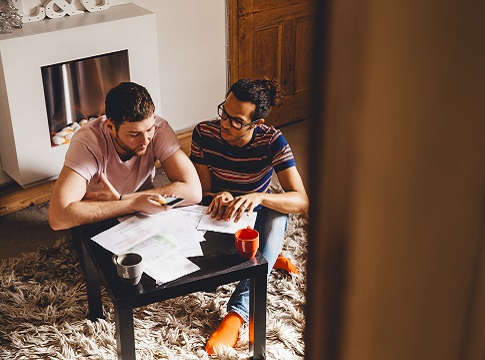 Male couple looking through financial documents together in their home. One of them is using his smartphone as a calculator which they are both looking at dissapointedly.